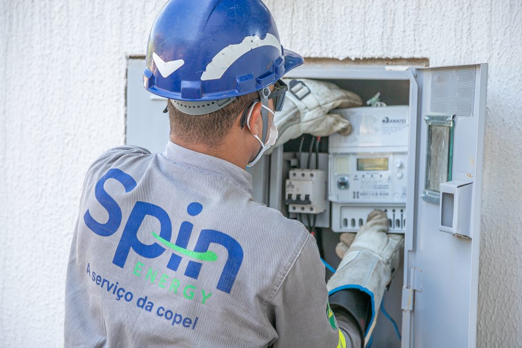 Homem uniformizado e de costas está diante de uma caixa de energia, instalando um novo medidor digital inteligente de energia