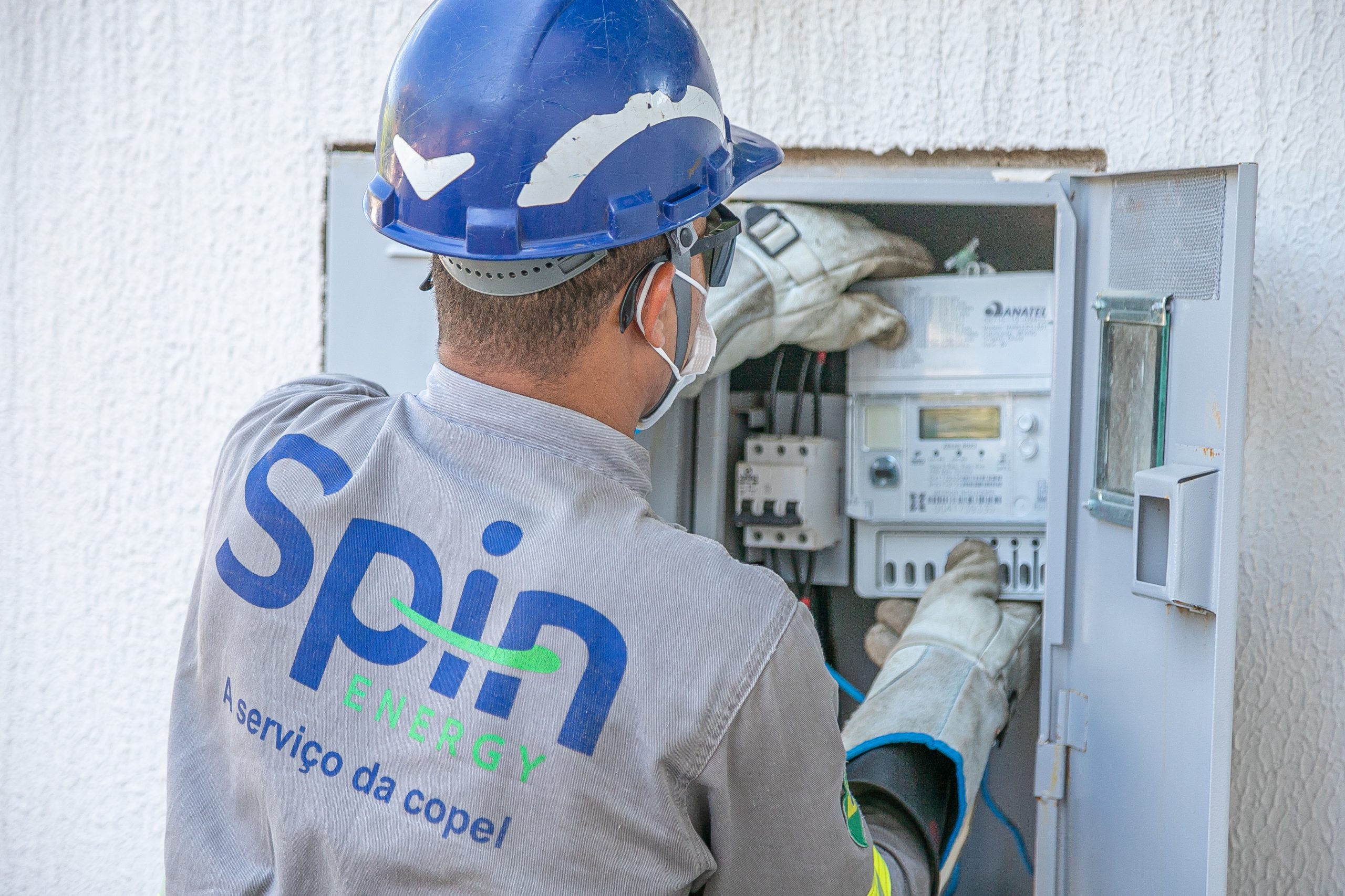 Homem uniformizado e de costas está diante de uma caixa de energia, instalando um novo medidor digital inteligente de energia