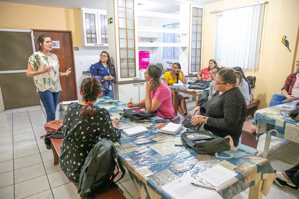 Mulher branca de blusa azul está em pé em frente a uma sala com aproximadamente seis mulheres sentadas às mesas para oficina de formação