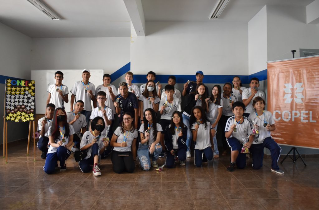 Turma grande de estudantes exibe medalhas conquistadas nas olimpíadas. À esquerda, um painel com 50 estrelas e os nomes dos medalhistas. À direita, um banner laranja escrito Copel