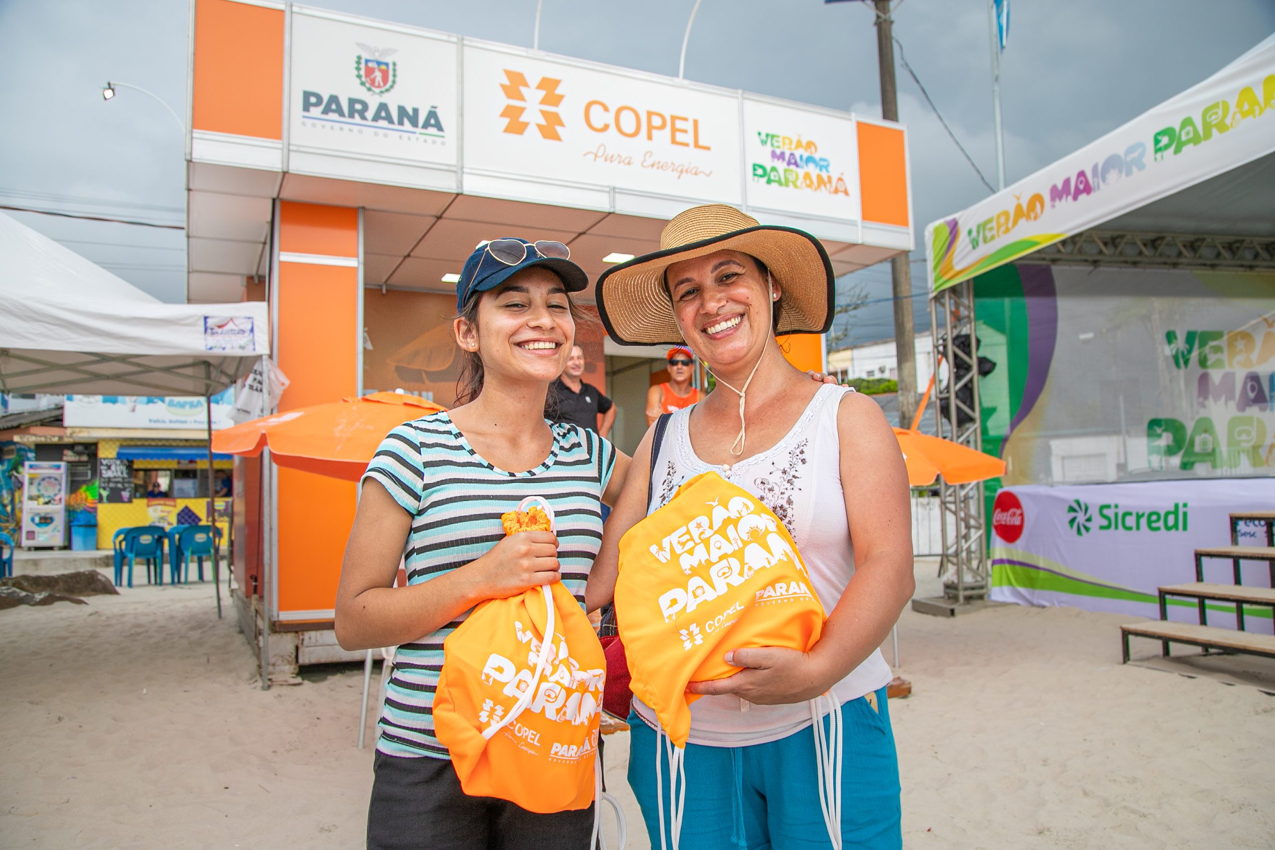 Uma adolescente e uma mulher em primeiro plano diante de estação da Copel na praia, segurando brindes