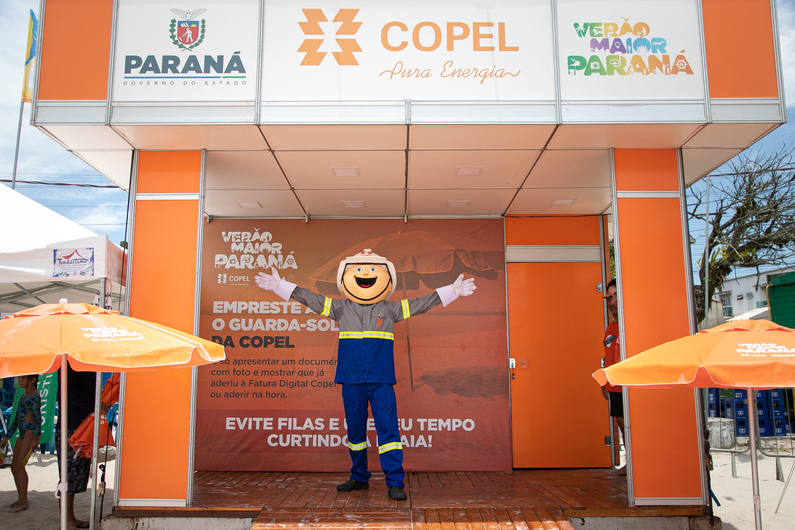 Pessoa vestida de Lucio, mascote da Copel, em frente à estação montada na orla para atividades recreativas