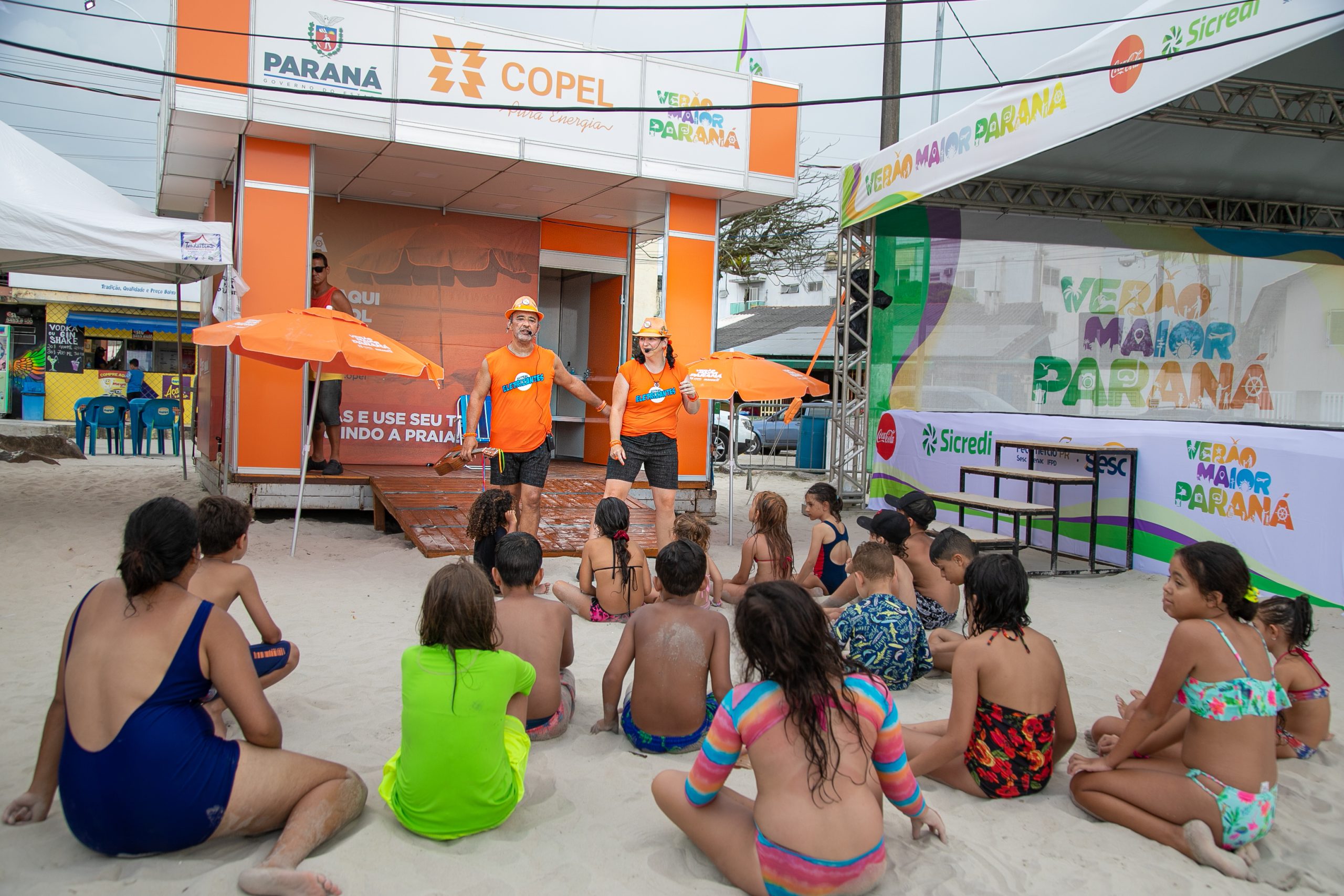 Atores da Copel apresentando peça teatral para um grupo de crianças sentadas na areia