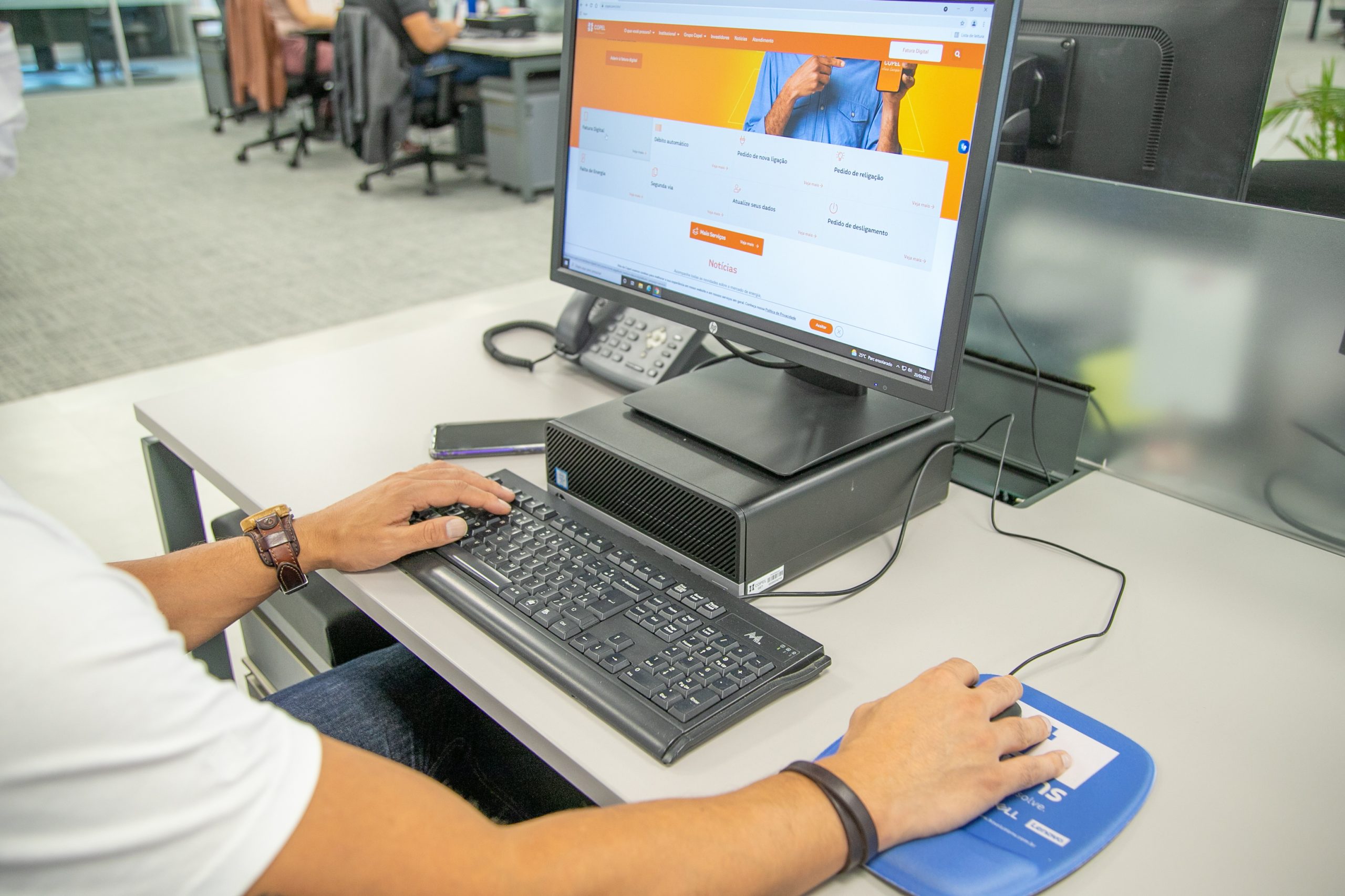 Pessoa acessa site da Copel em ambiente corporativo. Braços masculinos sobre a mesa com mão sobre mouse e teclado de computador