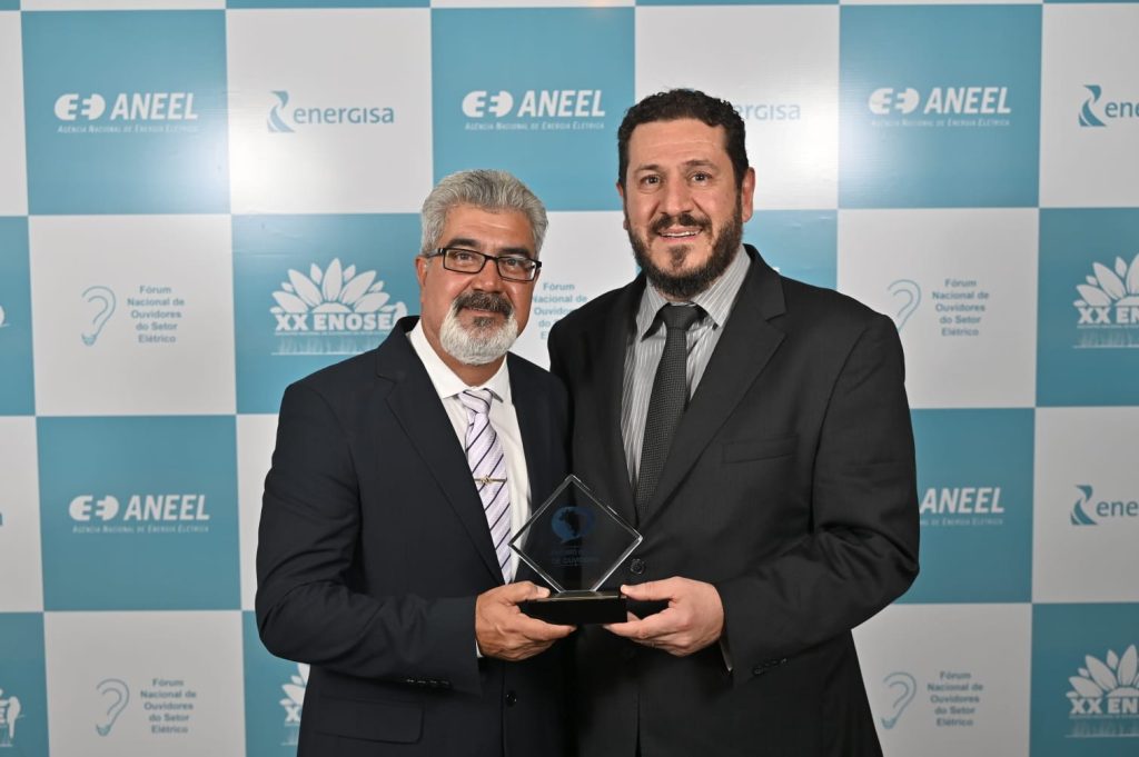 Ao fundo um painel com a logomarca da Aneel. À frente, Ubirajara Brum da Silva e Maximiliano Andres Orfali seguram o troféu recebido pela Copel. Estão de terno escuro e sorridentes