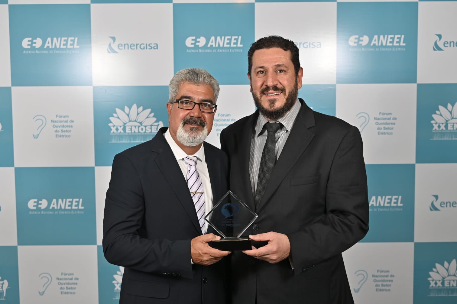Ao fundo um painel com a logomarca da Aneel. À frente, Ubirajara Brum da Silva e Maximiliano Andres Orfali seguram o troféu recebido pela Copel. Estão de terno escuro e sorridentes