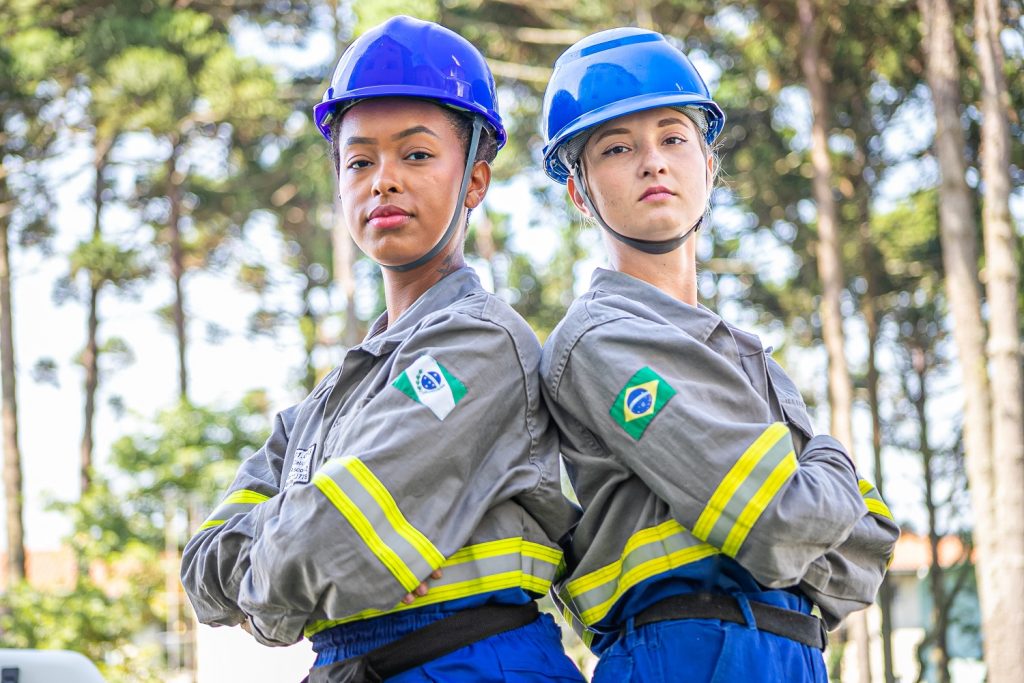 Mulheres eletricistas de uniforme semelhante ao da Copel e capacete azul, de braços cruzados com árvores ao fundo. À esquerda, uma mulher morena, à direita, uma mulher loira.