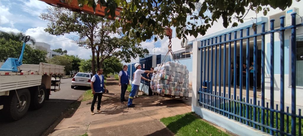 Caminhão com guincho segura pallet cheio de alimentos no portão de uma instituição beneficente. Três voluntários acompanham a movimentação do material.