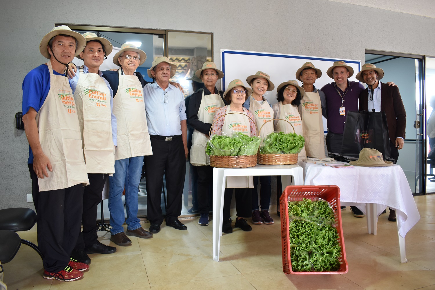 Voluntários da horta estão com avental e chapéu com a logomarca Cultivar Energia. O grupo está em pé, e à frente estão duas cestas e uma caixa com pés de alface colhidas na horta do programa