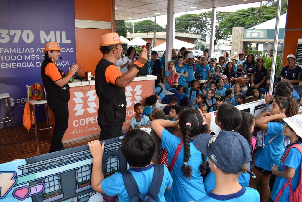 Educadores em pé à frente de uma grande turma de crianças com uniforme azul da rede municipal de ensino de Londrina, em um estande com laterais abertas