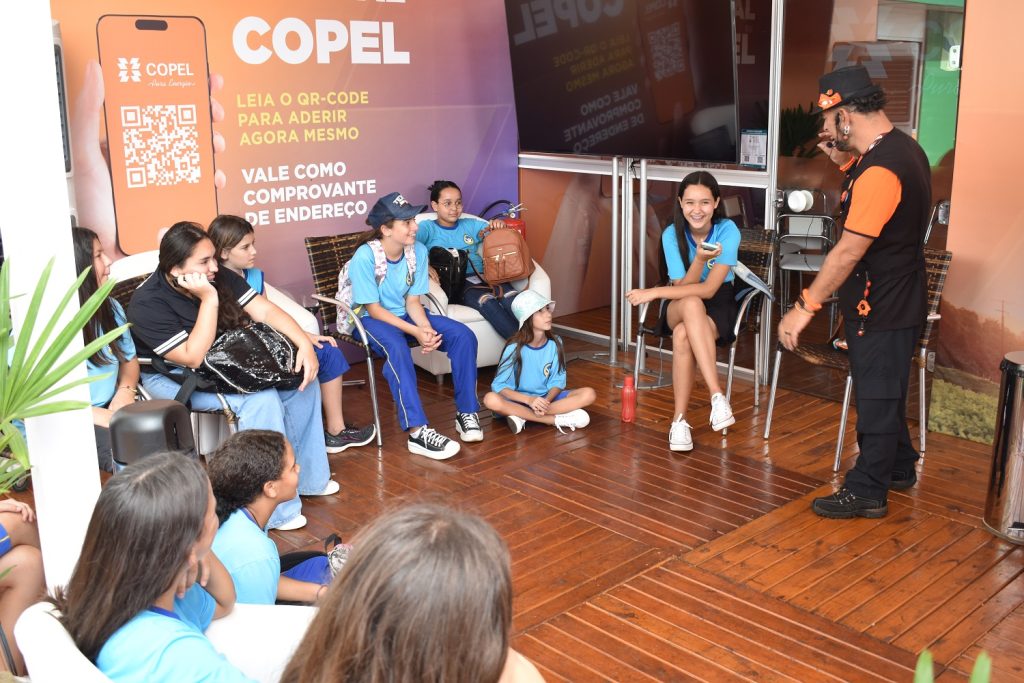 Alunos de uniforme azul estão sentados em círculo e o educador está em pé à frente deles, de camiseta laranja, calça e colete pretos, de chapéu na cabeça