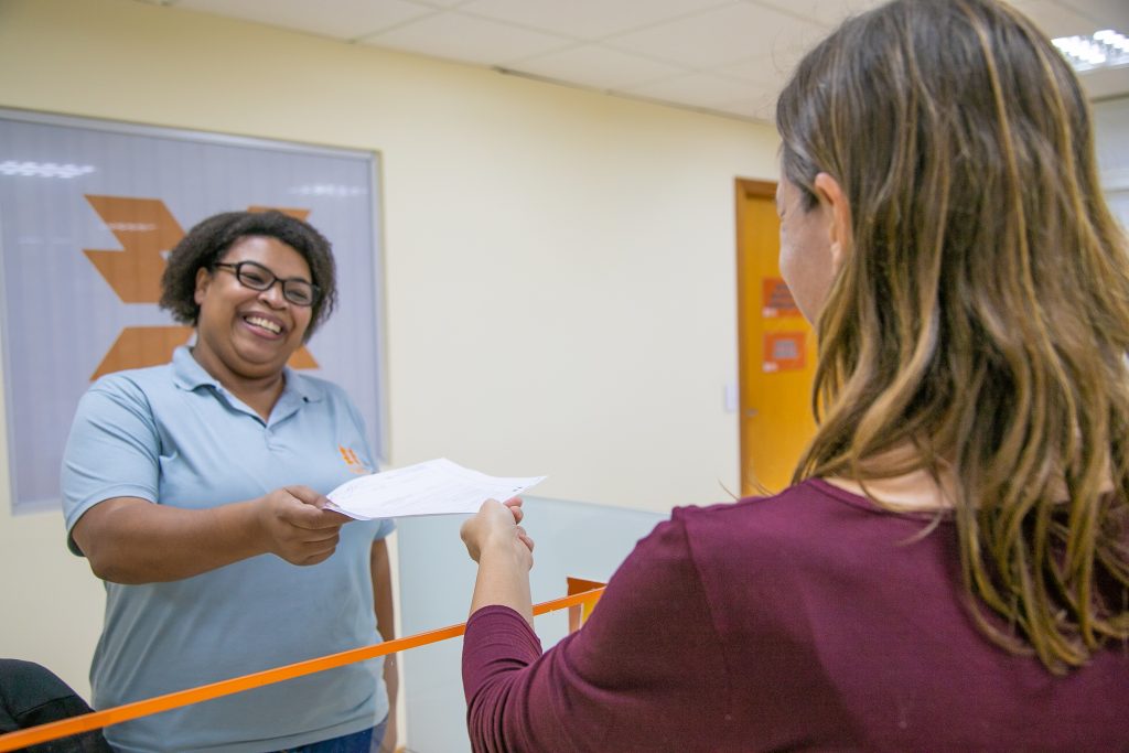 Atendente mulher negra sorri entregando papel para cliente mulher branca de costas. As duas estão em pé. Ao fundo a logomarca da Copel.