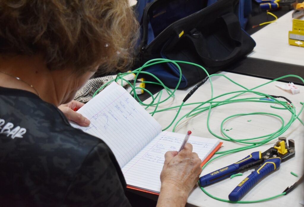 Mulher de costas faz anotações em caderno sobre mesa com alicate e fios