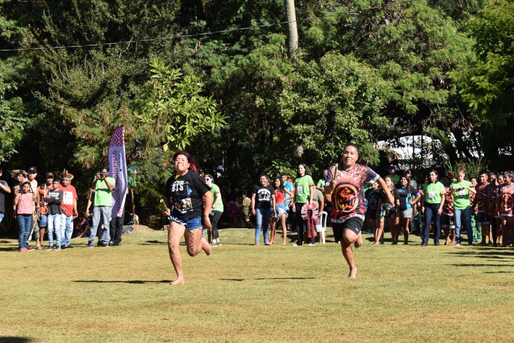 Mulheres indígenas disputam corrida em parque gramado com torcida e outros competidores ao fundo