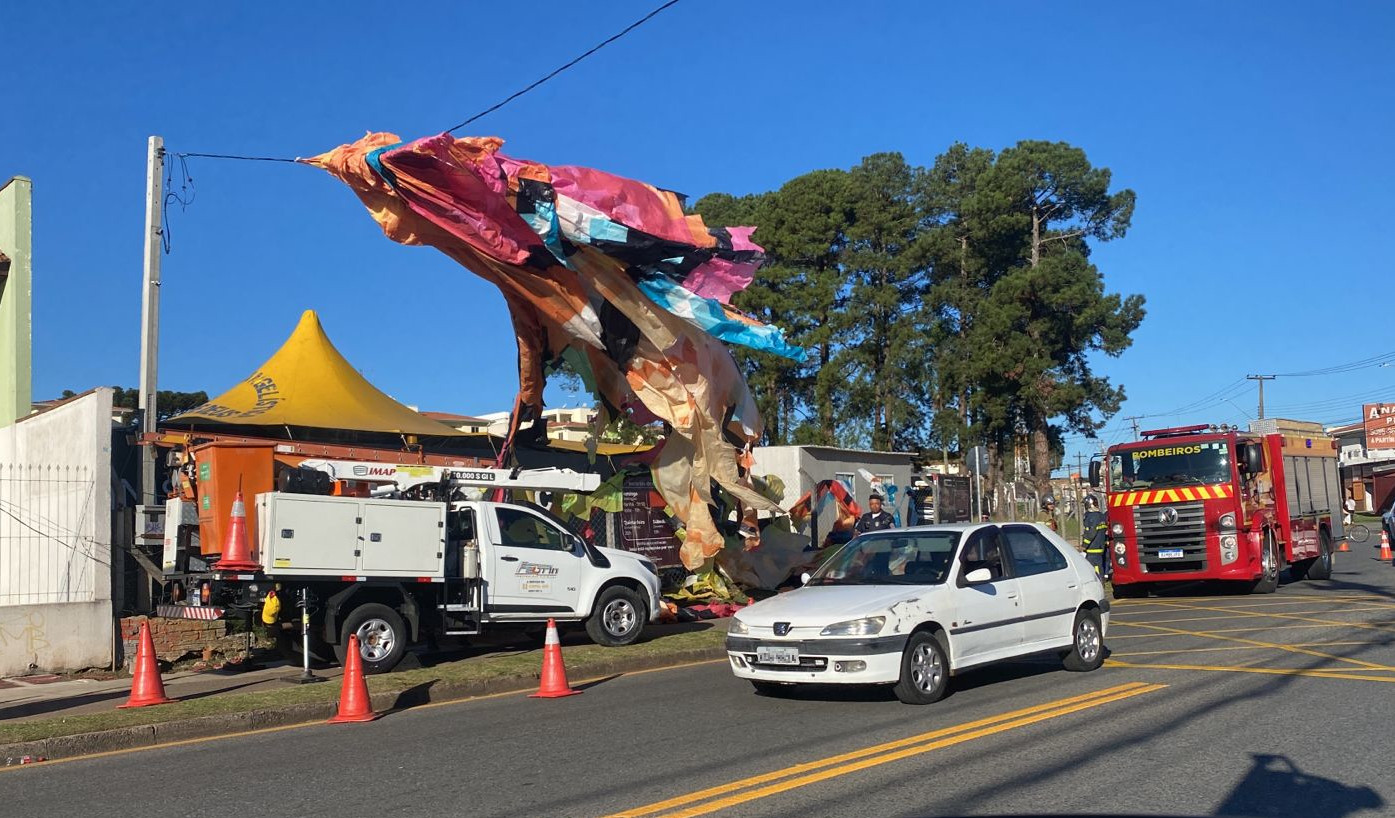 Balão de grande porte caído sobre a rede elétrica em rua com carros passando e caminhão do Corpo de Bombeiros