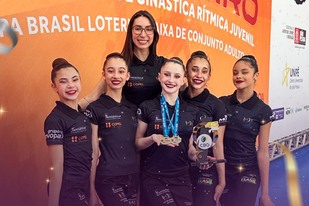 Cinco atletas sorridentes com a treinadora em frente a um painel da competição de ginástica rítmica