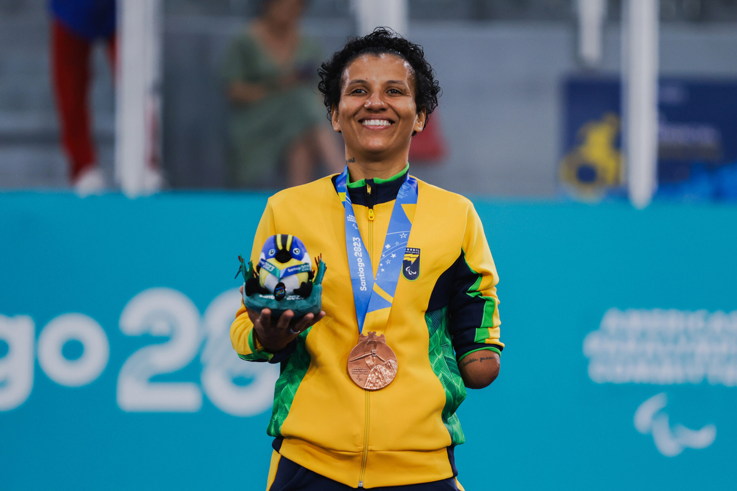 Atleta de jaqueta amarela com deficiência no braço esquerdo, segura mascote dos jogos na mão direita e leva no peito a medalha dos jogos parapanamericanos