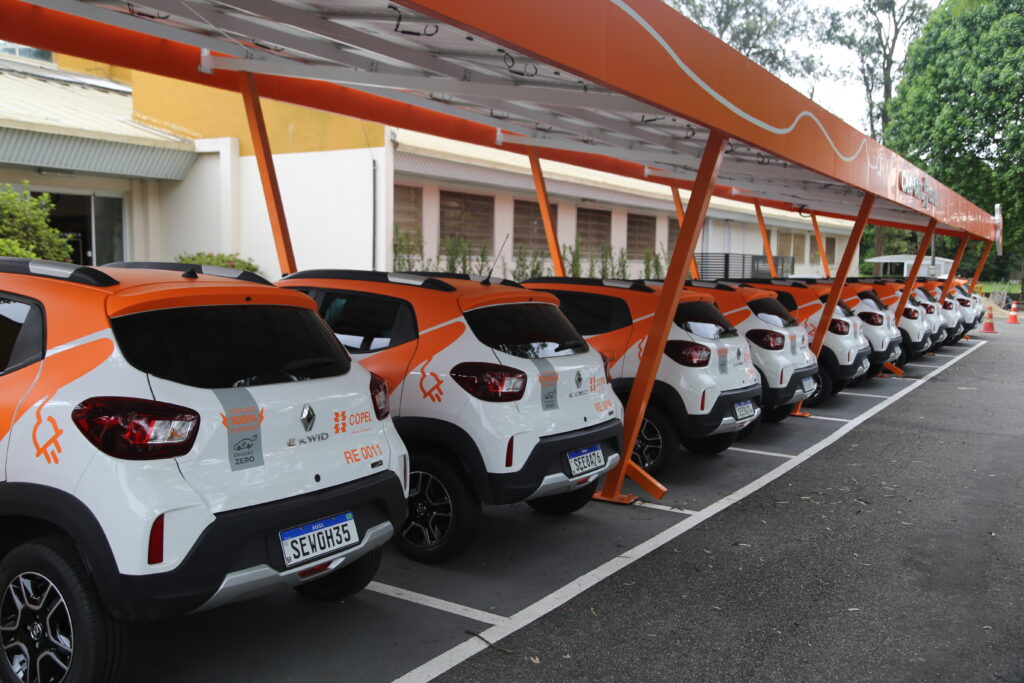 Fileira de veículos elétricos estacionados no pátio da Copel. Os carros são brancos e laranja.