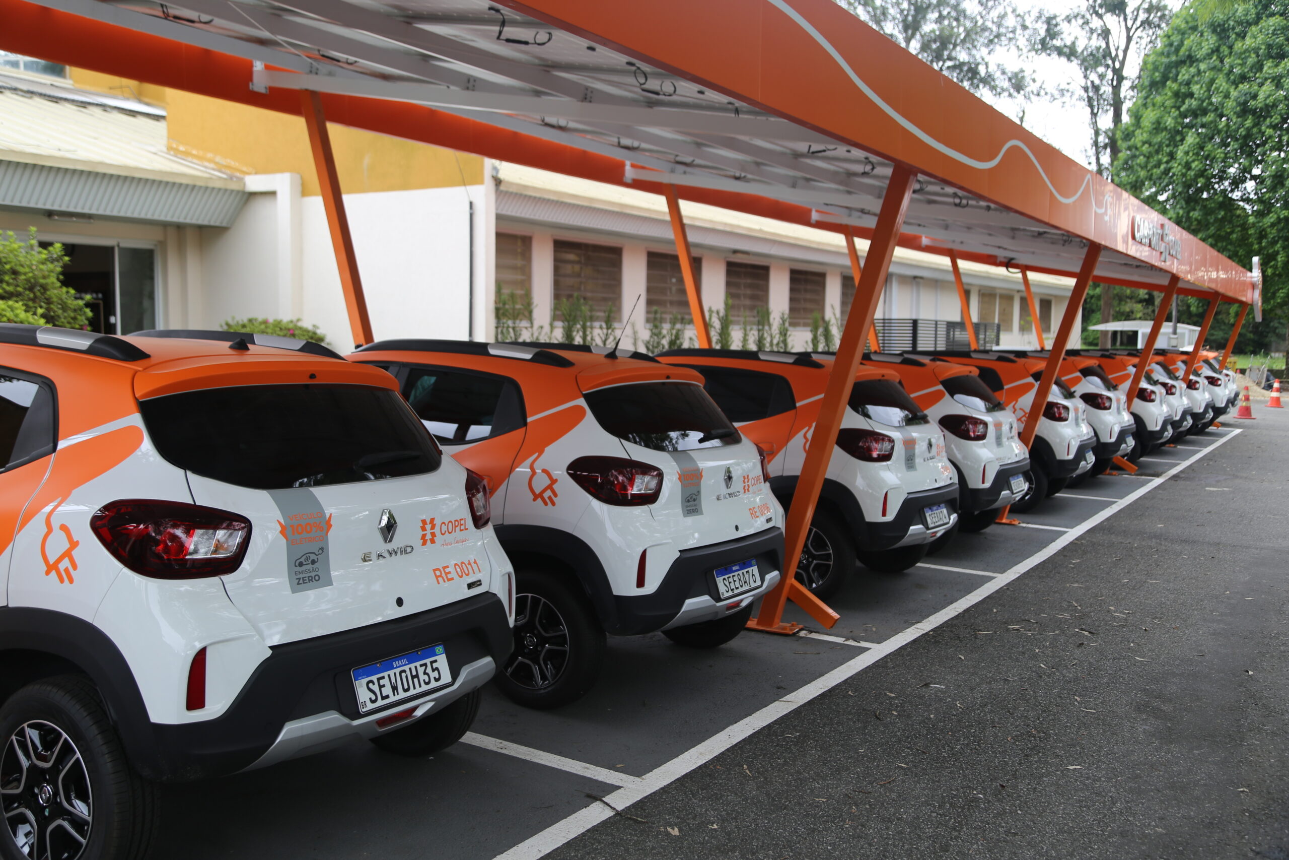 Fileira de veículos elétricos estacionados no pátio da Copel. Os carros são brancos e laranja.