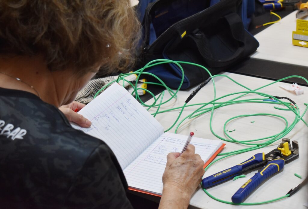 Mulher de costas escrevendo em um caderno. Sobre a mesa, alicate e fios elétricos.