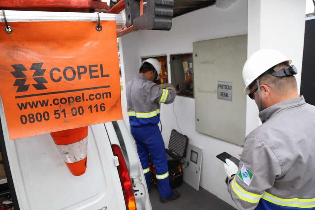 Dupla de eletricistas faz inspeção em um sistema de medição. Em primeiro plano, uma bandeirola de sinalização de veículo com a marca da Copel e o número de telefone 0800 51 00 116.