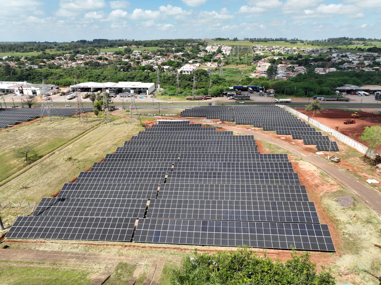 Imagem aérea de uma usina solar, com placas azuis em um campo gramado e parte da cidade ao fundo