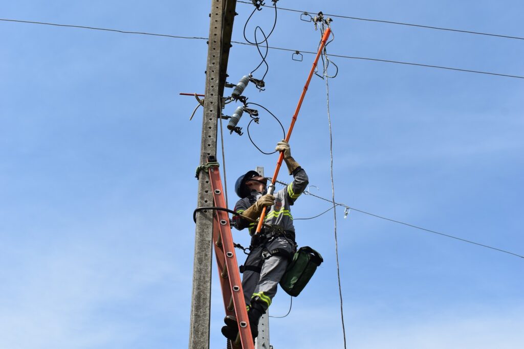 Eletricista sobre escada no alto do poste, de uniforme cinza, com um bastão laranja nas mãos