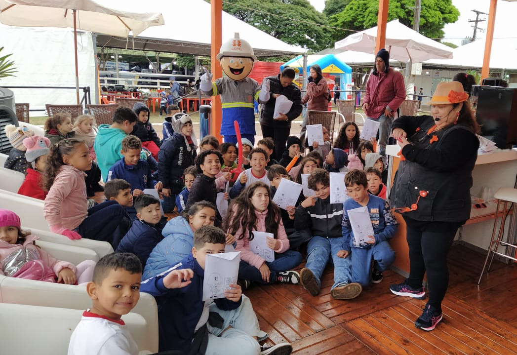 Estande da Copel com crianças reunidas, mascote Lucio e educadora em primeiro plano.