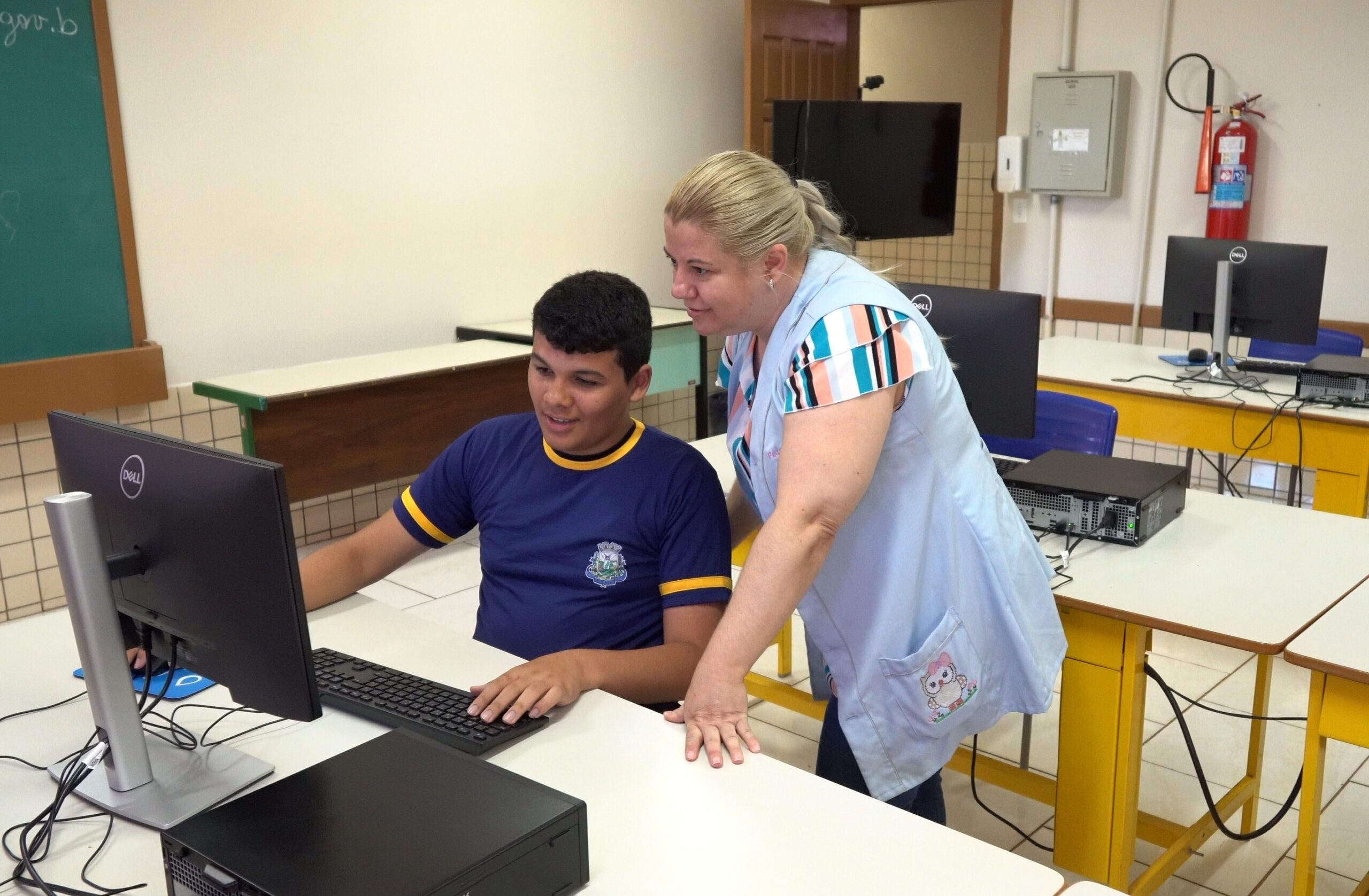 Aluno de uniforme azul marinho está sentado em frente ao computador e a professora ao lado em pé o auxilia