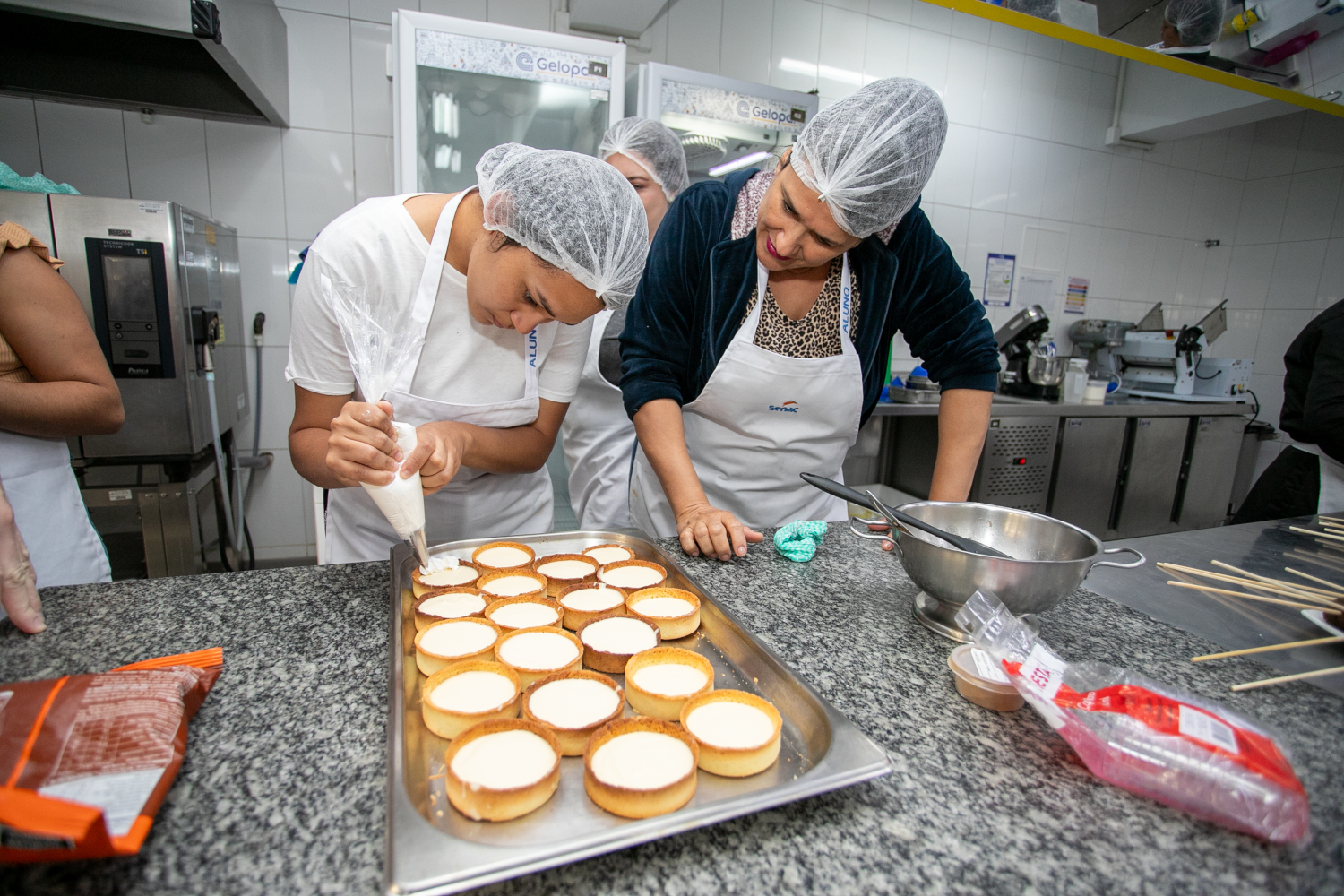 Duas mulheres de avental e toca no cabelo preenchem forminhas de confeitaria