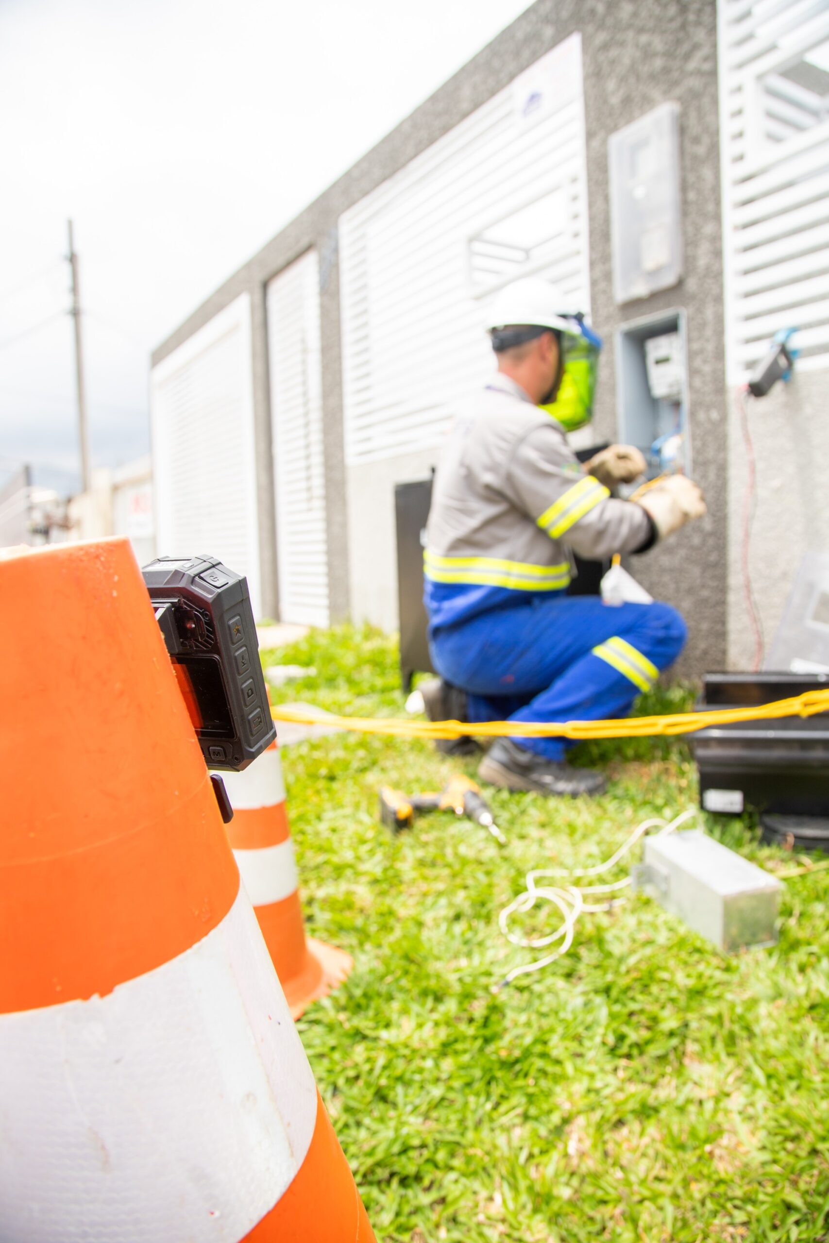 Em primeiro plano, uma câmera acoplada em um cone de sinalização acompanha o trabalho. Em segundo plano, homem eletricista uniformizado e com capacete e protetor facial consta de joelhos diante de caixa de energia trocando medidor de energia.